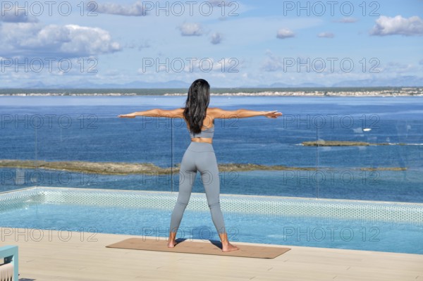 A woman stands on a yoga mat by an infinity pool, facing the ocean, arms extended in a peaceful yoga pose. The clear sky and serene sea create a calming backdrop