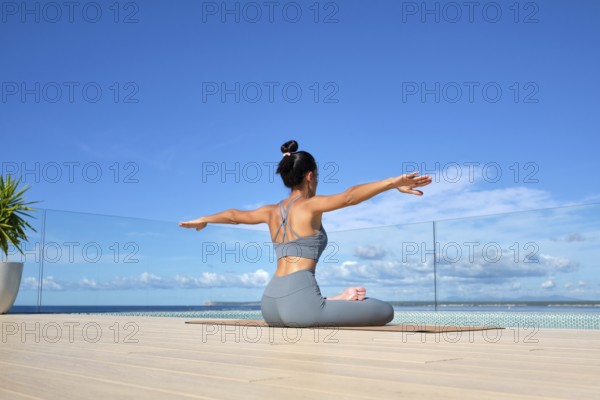 A woman in a gray yoga outfit practices yoga on a deck overlooking the ocean. Her arms are outstretched as she sits, embracing the tranquil blue sky and sea