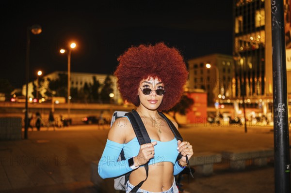 An African descendent woman with vibrant afro hair stands confidently on a city street at night. Dressed in casual, spring wear, she sports sunglasses and carries a backpack
