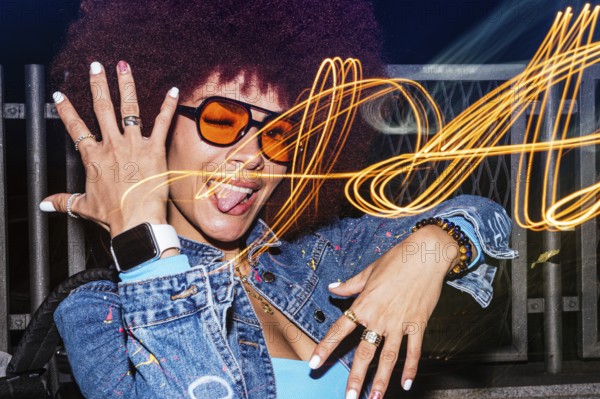 African descendant woman with afro hair showcases her playful side, winking and sticking her tongue out with orange-tinted sunglasses at night. Light trails swirl in the background, adding a dynamic and whimsical atmosphere