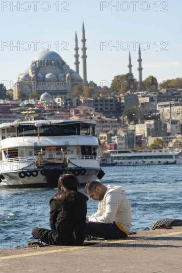 Istanbul, Turkey 10th November 2022 Daily life in Istanbul during an economic crisis, a couple sit beside the Golden Horn with a ferry and mosque in the background
