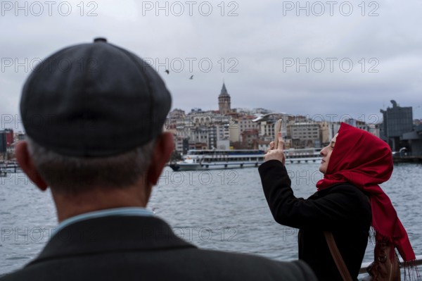 Istanbul, Turkey, February 23rd 2018. A Turkish Muslim woman taking photographs on the Golden Horn with the Galata tower in the background, the European side of Istanbul, Turkey