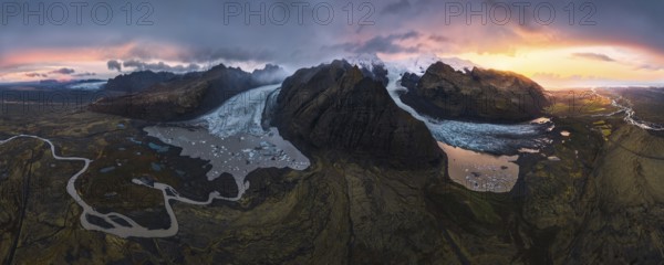 A stunning aerial view of a glacier cascading between volcanic mountains, surrounded by glacial lakes, winding rivers, and dramatic skies illuminated by a vibrant sunset in Iceland