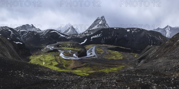 A striking view of black volcanic mountains partially covered in snow, with a winding river and vibrant green moss in the valley, under a dramatic and moody sky in Iceland's highlands