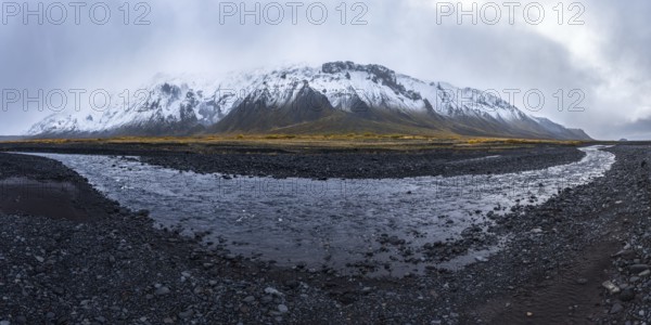 A dramatic view of a snow-covered mountain rising over a rocky riverbed in the Icelandic Highlands, featuring dark volcanic terrain and a moody overcast sky