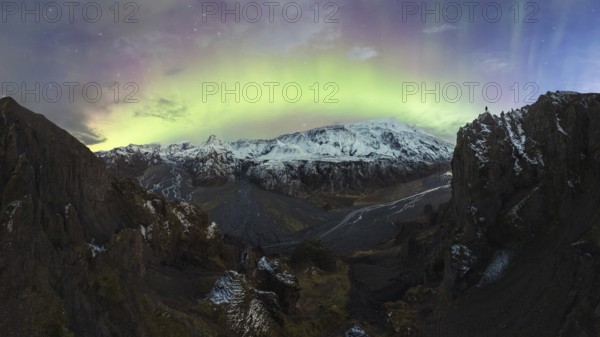 Breathtaking view of the Northern Lights illuminating the sky above a snow-covered mountain range and rugged terrain in Iceland