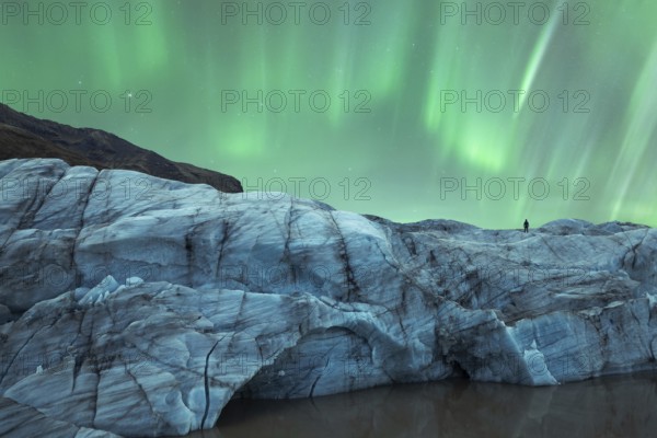 A solitary figure stands atop a glacier in Iceland, gazing at the mesmerizing Northern Lights that illuminate the night sky