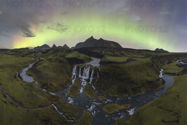 The aurora borealis illuminates the sky above a stunning Icelandic waterfall and meandering river surrounded by rugged terrain