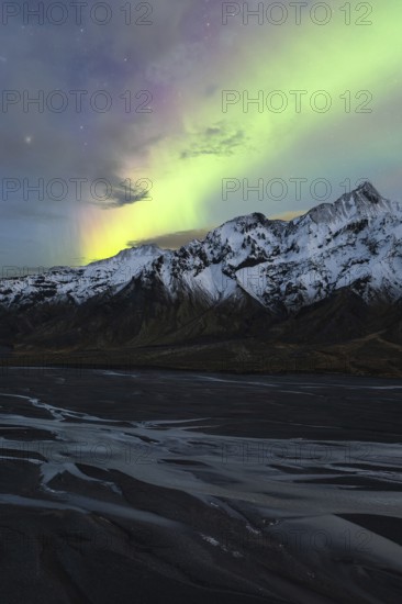 Captivating Northern Lights (Aurora Borealis) shine above snow-covered peaks and a riverbed in the Icelandic wilderness, creating a stunning natural spectacle