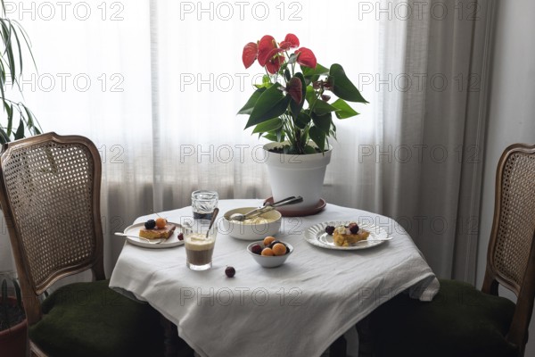 Warmly lit coffee table covered in a white cloth, featuring various desserts like cherry-topped cakes and ice cream, a potted red-flowering plant, coffee, and refreshing water, presenting an inviting and peaceful atmosphere