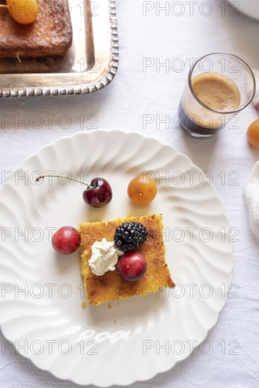 Top view of a gourmet desert scene on a light tablecloth, featuring a piece of cake topped with cream and fresh berries, accompanied by cherries, a small apricot, and a shot of espresso