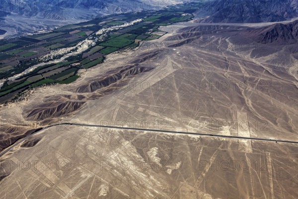 Aerial photo shows dry geoglyphic landscape next to green fields and a road, the geoglyphs and drawings in the desert near Nasca and Palpa in Peru
