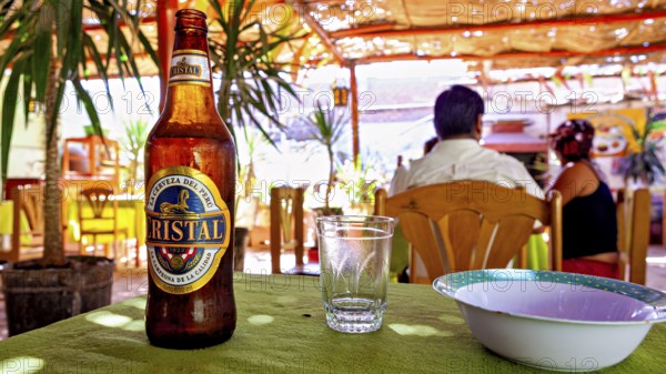 Scene in a bright restaurant with beer bottle and glass on a table, guests and tropical plants in the background, the city of Nasca in Peru