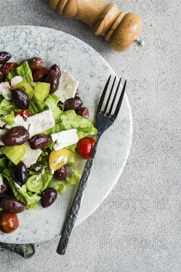 A vibrant Greek salad with tomatoes, lettuce, red olives, and feta cheese Complemented by a pepper shaker, all presented on a textured table