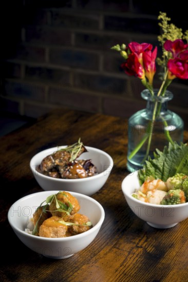 A styled display featuring tempura mushrooms in curry sauce and aubergine stew served over rice Arranged in elegant white bowls on a rustic wooden table with floral decoration in the background