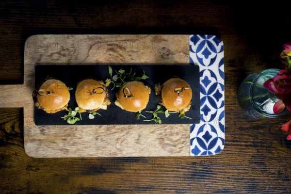 Top-down view of four gourmet pulled pork and cabbage mini burgers served on a black slate board with fresh greens, arranged over a rustic wooden tray Perfect for appetizers or finger food