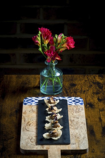 Elegant cracker canapes topped with vegetable satay stew and peanuts, served on a black slate over a wooden board A vase with red flowers adds a vibrant touch to the rustic dining setup