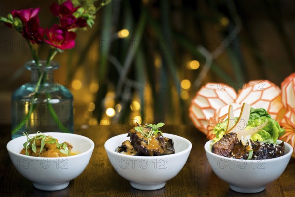 Trio of gourmet dishes, tempura mushrooms in curry sauce, aubergine stew over rice, and roasted beef with greens, pear, and sesame seeds The festive setting features warm lighting and floral decor