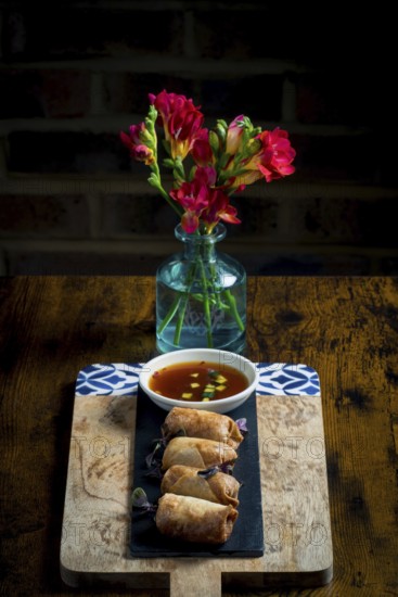 A wooden board holds crispy spring rolls beside a bowl of dipping sauce, set against a vibrant bouquet of red flowers in a clear vase, creating a cozy dining setting