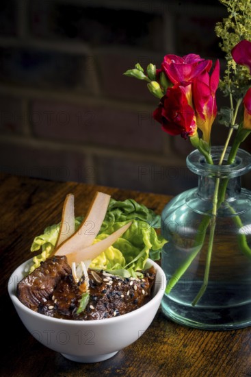 A beautifully presented Asian beef bowl garnished with fresh greens and pear slices, paired with a vibrant bouquet in a glass vase on a wooden table