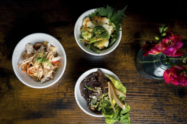 Top-down view of three gourmet bowls, broccoli and cauliflower in satay sauce, chicken with glass noodles and peanuts, and roasted beef with fresh greens, pear slices, and sesame seeds