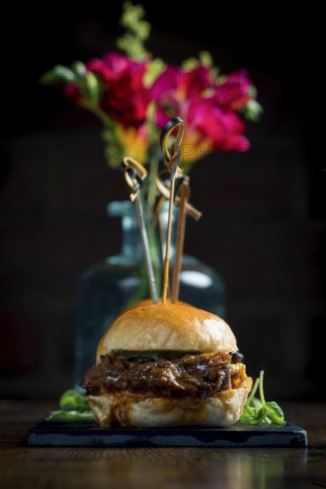 A gourmet pulled pork slider presented elegantly with colorful flowers in the background The food is garnished on a slate, creating an exquisite presentation