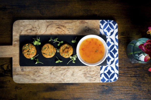 Top-down view of golden corn and spring onion patties served with a bowl of sweet chili sauce, garnished with fresh greens Beautifully arranged on a slate board over a rustic wooden table
