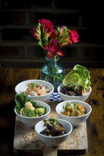 A selection of vibrant Asian cuisine bowls arranged on a wooden board The dishes feature vegetables, meats, and garnishes A vase of bright red flowers adds a decorative touch in the background