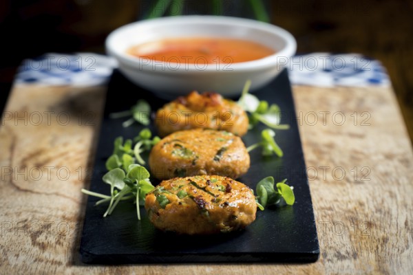 Close-up of golden corn and spring onion patties served with fresh greens and accompanied by a bowl of sweet chili sauce The dish is elegantly presented on a slate board with a rustic backdrop