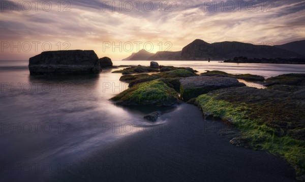 Sunrise casts golden hues over the rocky shoreline of Cala del Lastre in Portman, Murcia The tranquil sea contrasts with the rugged moss-covered rocks