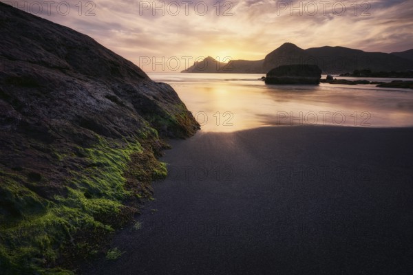 The tranquil sunset casts a golden hue over the rugged rocks and calm waters of Cala del Lastre in Murcia, Spain, creating a peaceful and scenic coastal view