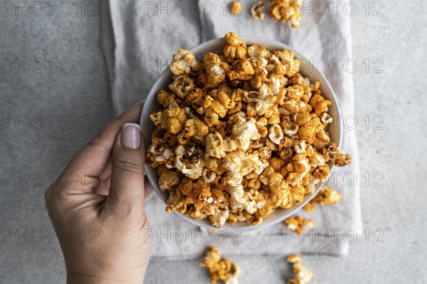 Cropped unrecognizable person holding a bowl of sweet caramel popcorn in a top view, displaying the texture and color of the snack against a muted gray background