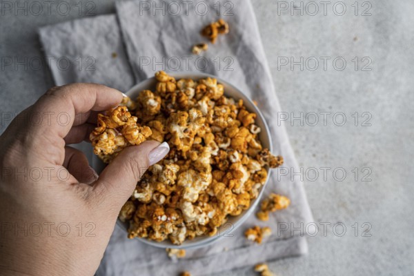 Cropped unrecognizable person's hand picking sweet caramel popcorn from a gray bowl, set against a soft gray background