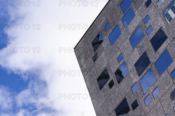From below view of a corner of a modern textured building facade featuring multiple aligned blue glass windows, set against a bright sky with soft clouds