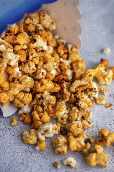 Caramel-coated popcorn spills from a blue container onto a textured gray surface, showcasing the crunchy treat's varied golden hues and textured pieces