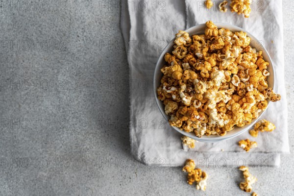 Top view of a bowl full of sweet caramel popcorn, sprinkled with sugar and spices, on a textured gray surface with loose kernels scattered around