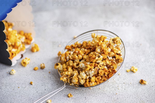 Top view of sweet caramel popcorn served in a transparent bowl on a textured gray background. The delicious snack is scattered around with focus on its rich, golden texture
