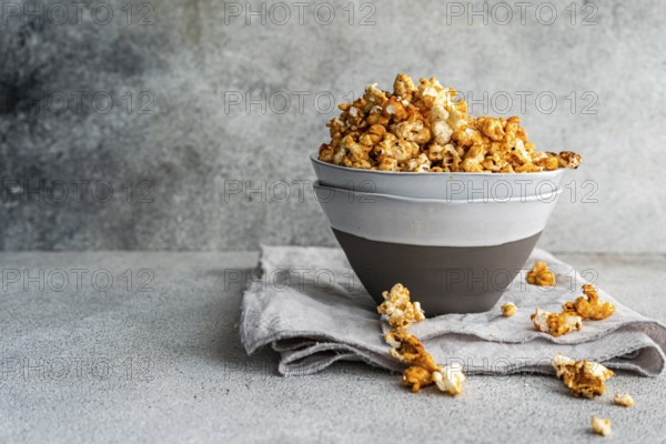 Gray bowl overflowing with sweet caramel popcorn is set on a textured linen, against a textured gray background to highlight the rich, golden tones of the snack