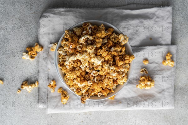 Top view of a bowl filled with sweet caramel popcorn, garnished with scattered pieces on grey textile background