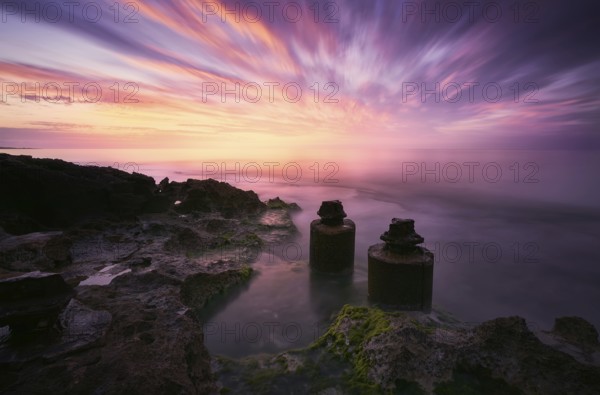 A mesmerizing sunset with vibrant hues reflecting over calm waters, coastal rocks, and ancient ruins or structures, creating a tranquil and dramatic seascape in Spain