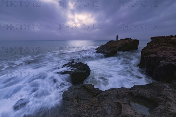 A dramatic seascape view at Cabo Cervera, Torrevieja, Alicante A lone figure stands on rocks, overlooking waves crashing against the rugged coastline under a moody sky
