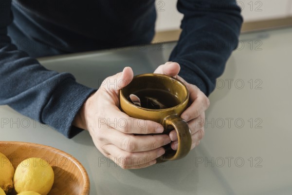 A close-up of hands holding a brown mug of tea with a spoon inside, placed on a glass table A wooden bowl with lemons adds warmth to the cozy and calm atmosphere