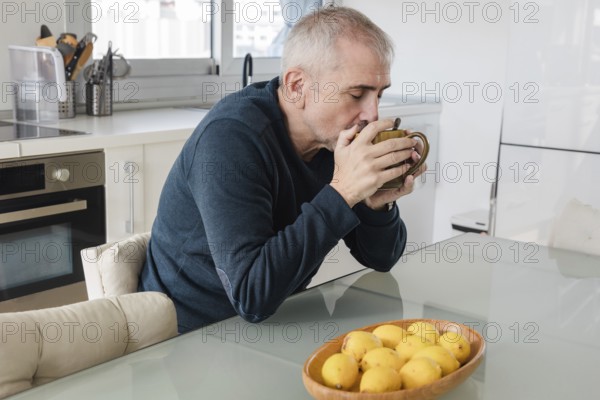 A senior man enjoys a warm cup of coffee at a glass table in a modern kitchen A bowl of lemons decorates the table, adding a pop of color to the serene setting