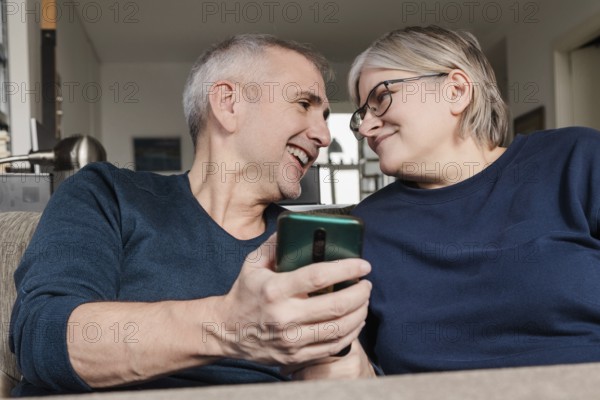 Smiling senior couple sitting on a couch, sharing a smartphone moment at home, demonstrating warmth, affection, and digital connectivity in their everyday life
