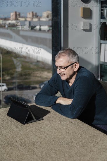 A senior man wearing glasses smiles as he engages with a tablet indoors Sunlight filters through the window, casting a warm ambiance in the room