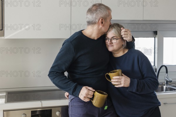 A senior couple shares a warm, affectionate moment in their kitchen, each holding a mug The scene captures the comfort and intimacy of daily life together