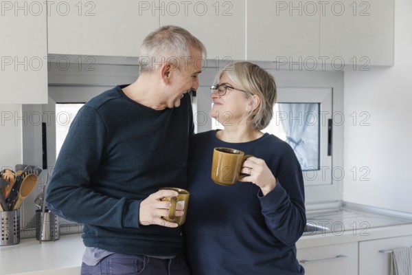 Senior couple shares a joyful moment in a modern kitchen, each holding a coffee cup Their warmth and connection create an atmosphere of comfort and companionship
