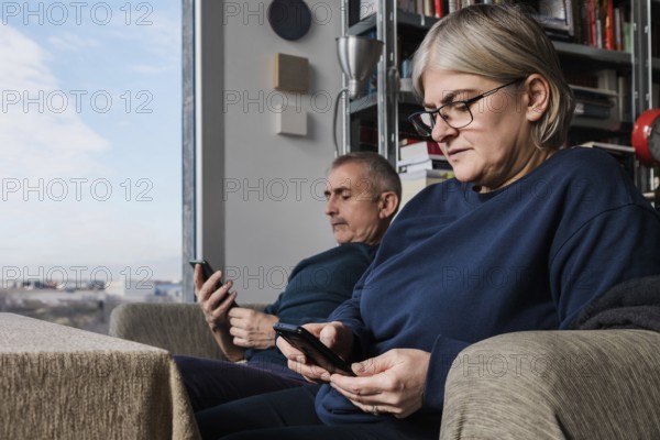 A senior couple sits in their cozy living room, each engaged with a smartphone The image captures modern connectivity and relaxation in a well-lit space with a city view