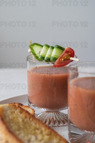 Clear glasses of gazpacho garnished with cucumber and tomato skewers A slice of crispy toast is served beside the glasses, all set on a neutral background