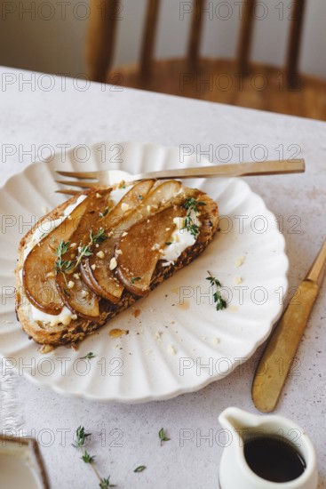 Top view of a delicious pear toast garnished with thyme, honey drippings, and crumbled cheese, served on a white scalloped plate alongside a small jug of honey and a wooden fork